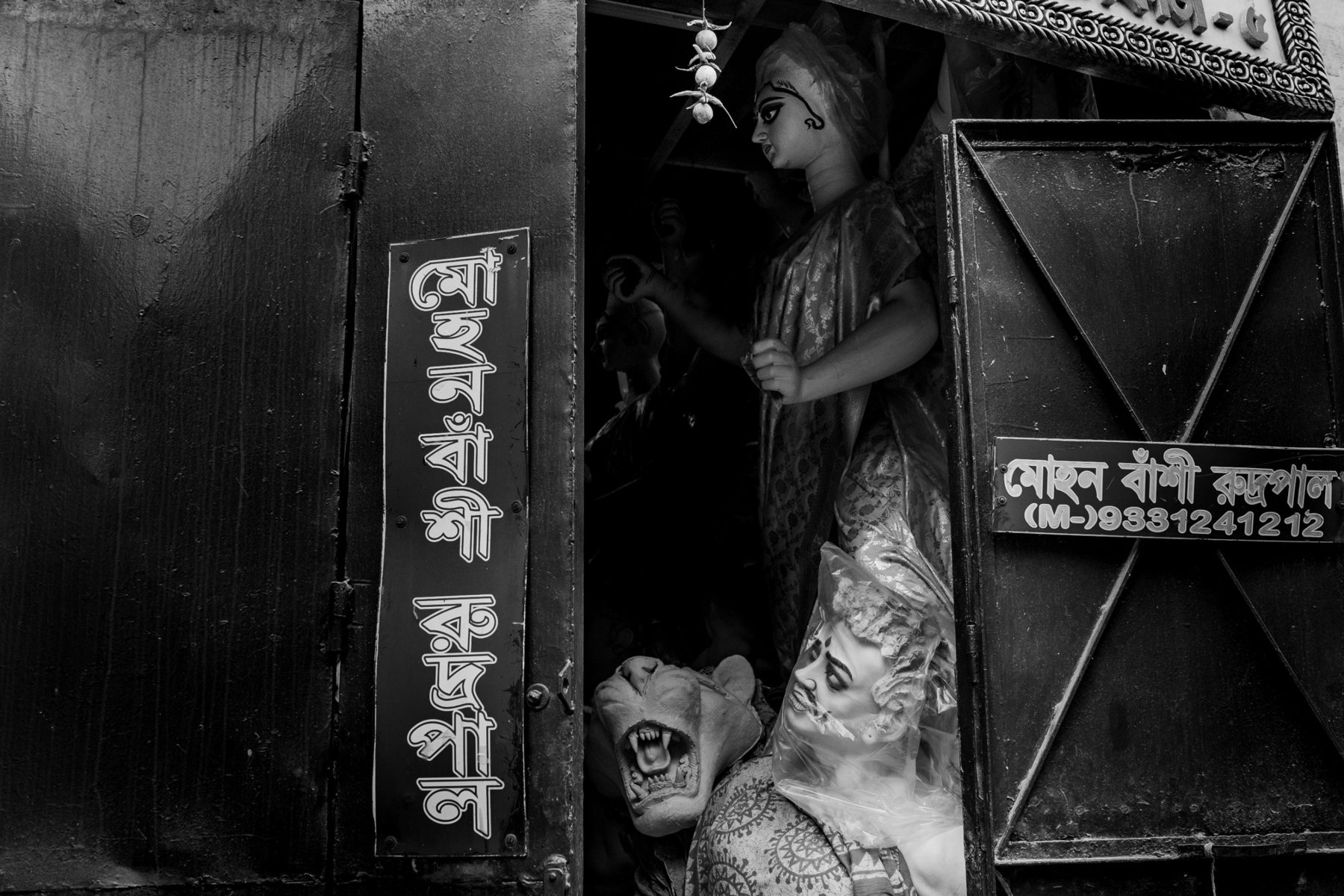A black workshop door in Kumartuli carrying a man's name in Bengali, painted vertically and on a plate with a phone number; inside, a goddess figure stands above a clay lion and a demon's head wrapped in plastic.