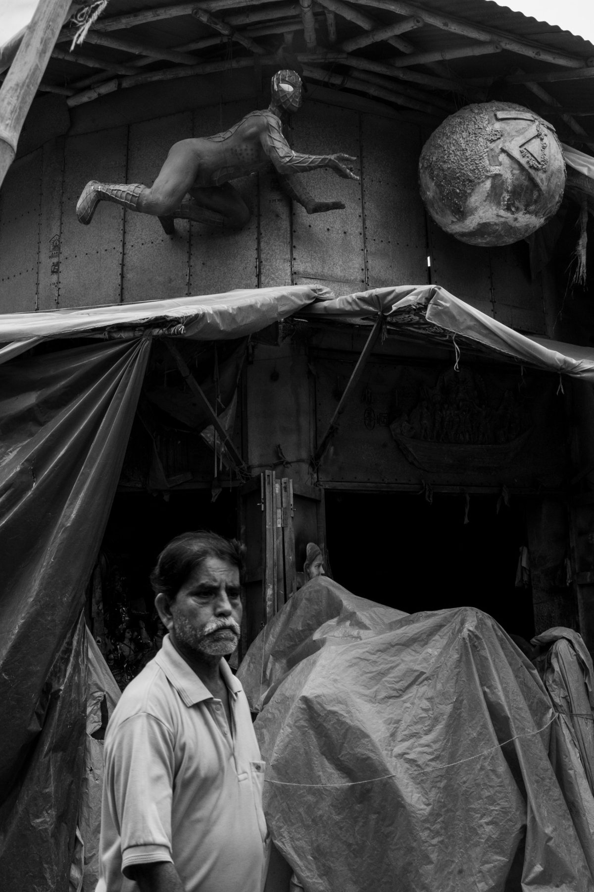 A grey-haired man walks past a workshop draped in tarpaulin, below a clay Spider-Man figure and a cracked globe fixed to the corrugated wall.
