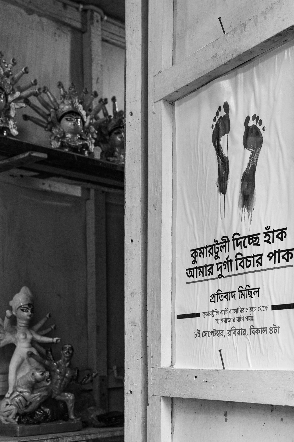 A protest poster with two bleeding footprints pinned to a white workshop door, beside shelves of small Durga figures.