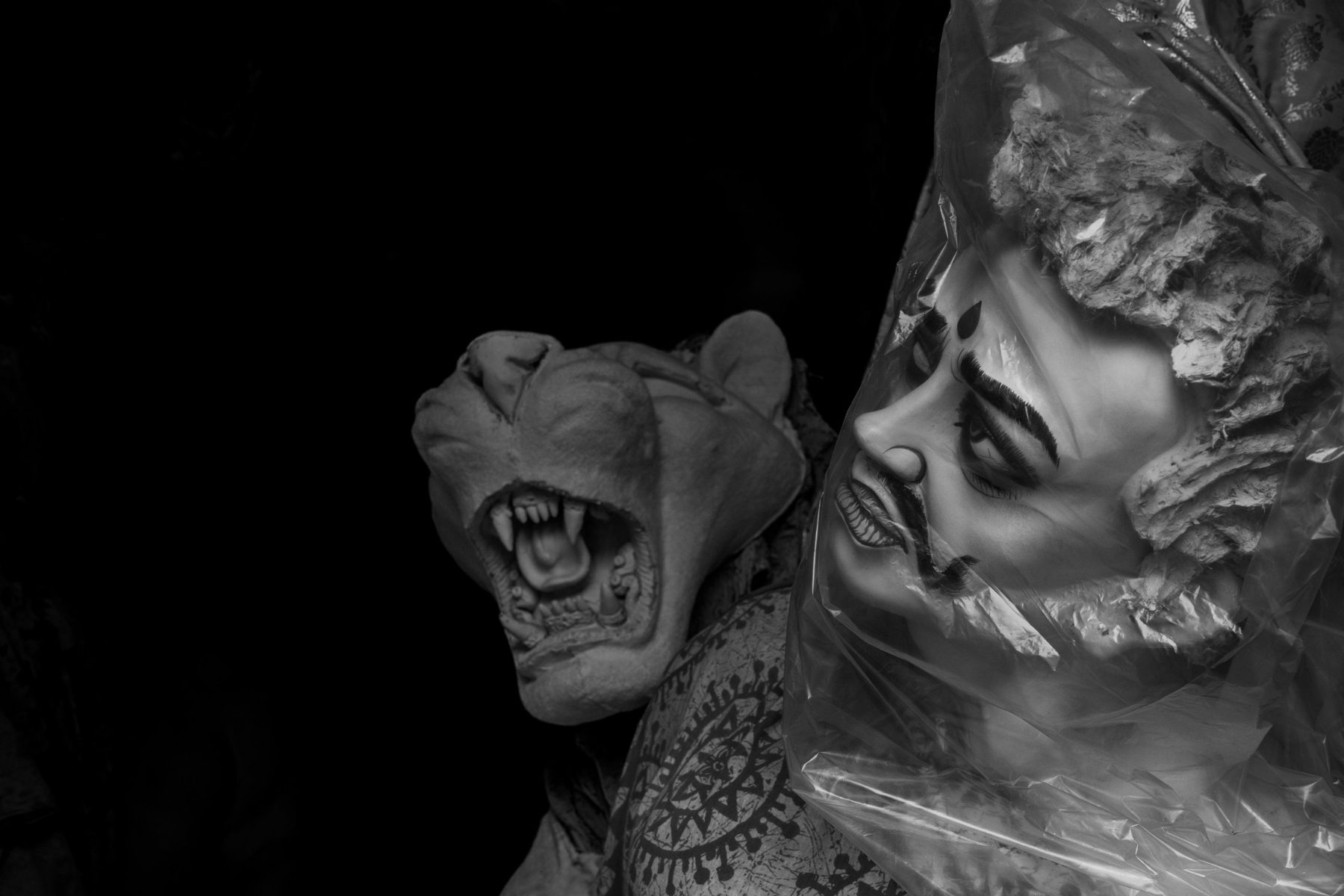 A snarling clay lion and the moustached head of the demon Mahishasura, wrapped in clear plastic, against black.