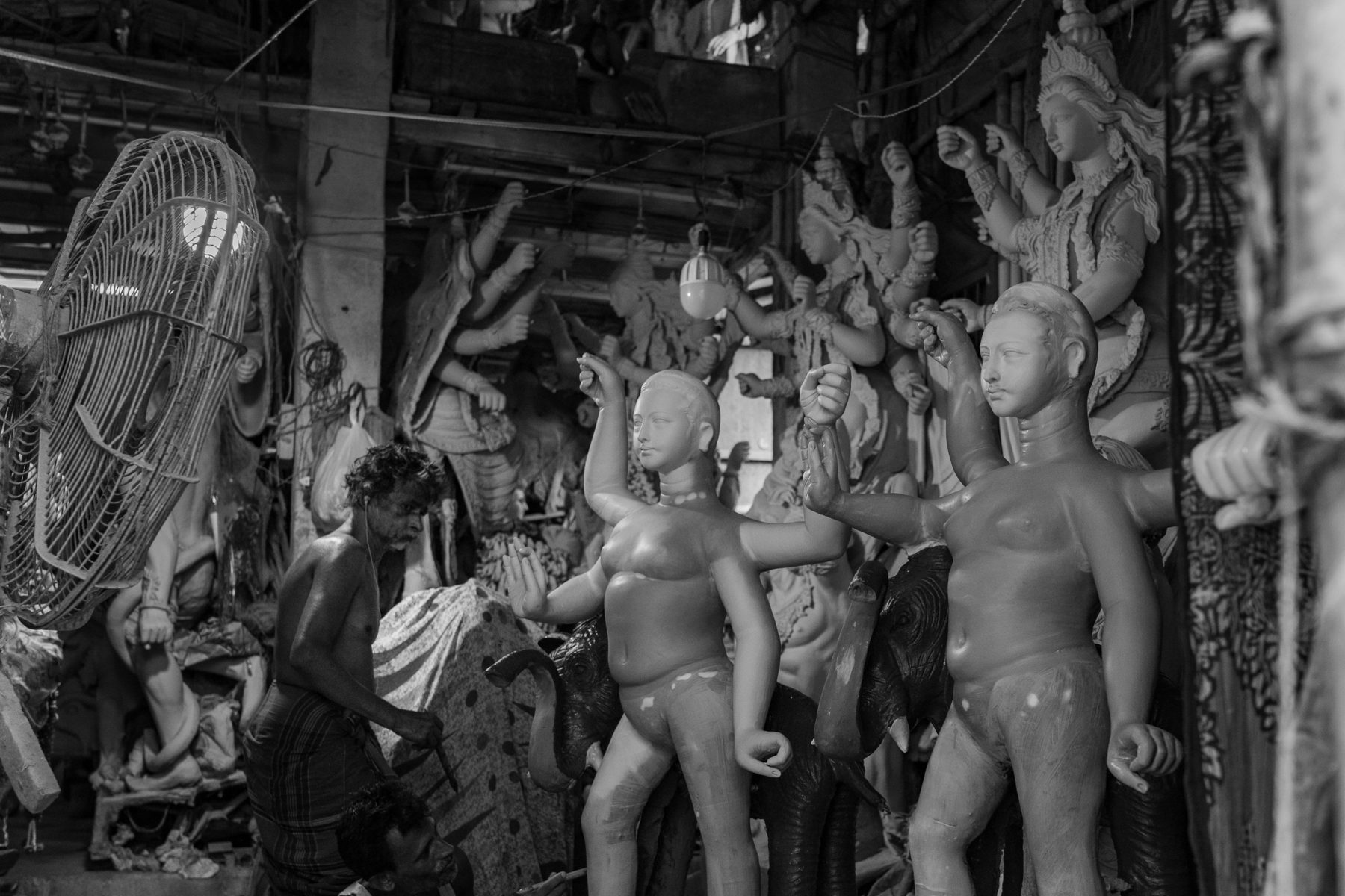 A bare-chested artisan works with a blade among unpainted clay goddesses with raised arms, an industrial fan and a hanging bulb beside them.