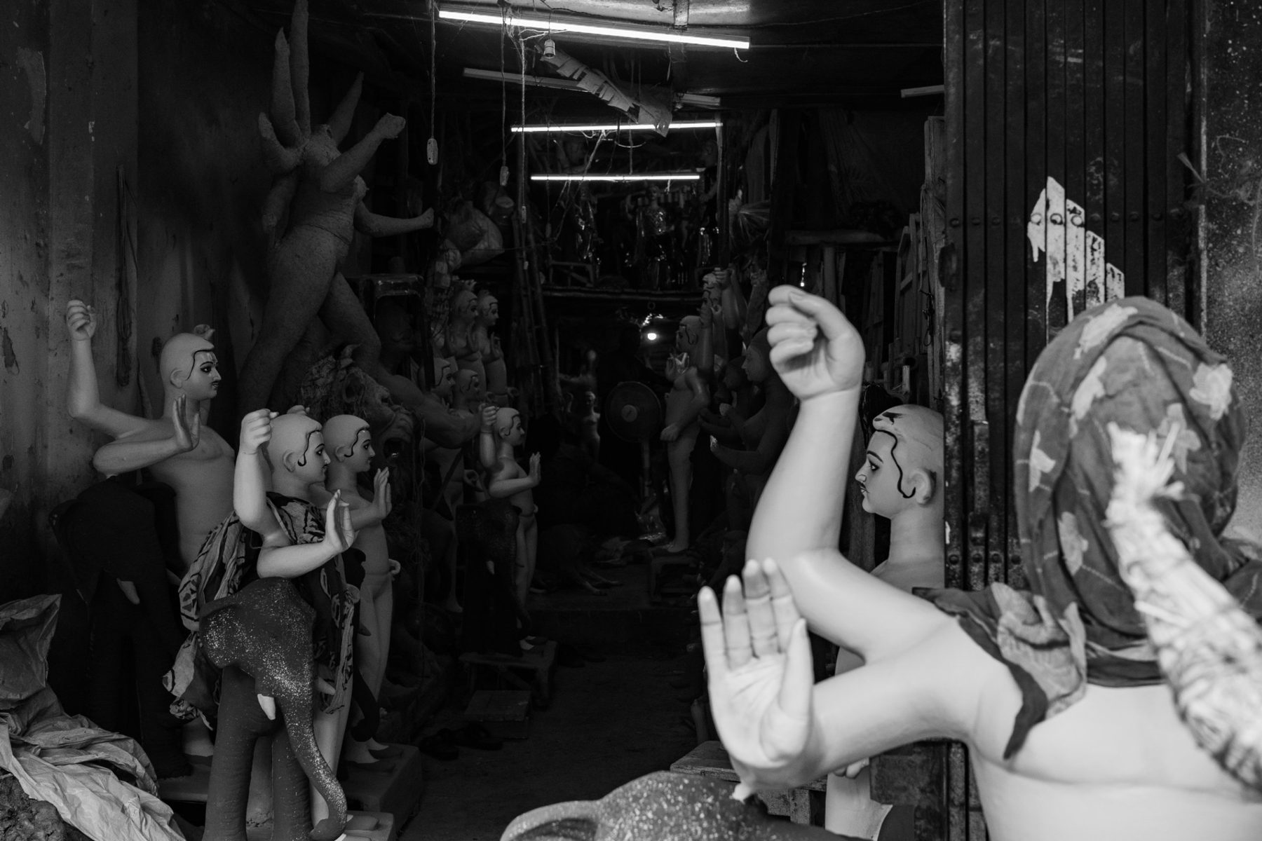 A long workshop corridor lined with unfinished figures, fists and palms raised; in the foreground a figure with a cloth over its head holds up a clenched fist.