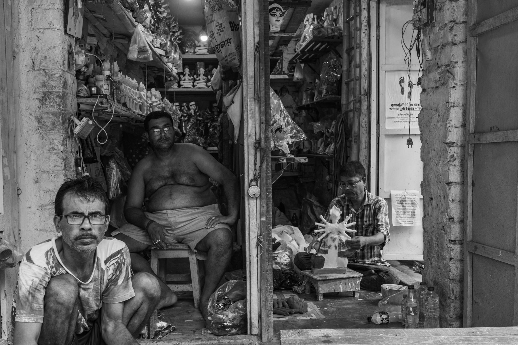 Three men in a Kumartuli workshop, one shaping a many-armed Durga, a protest poster on the door behind them.